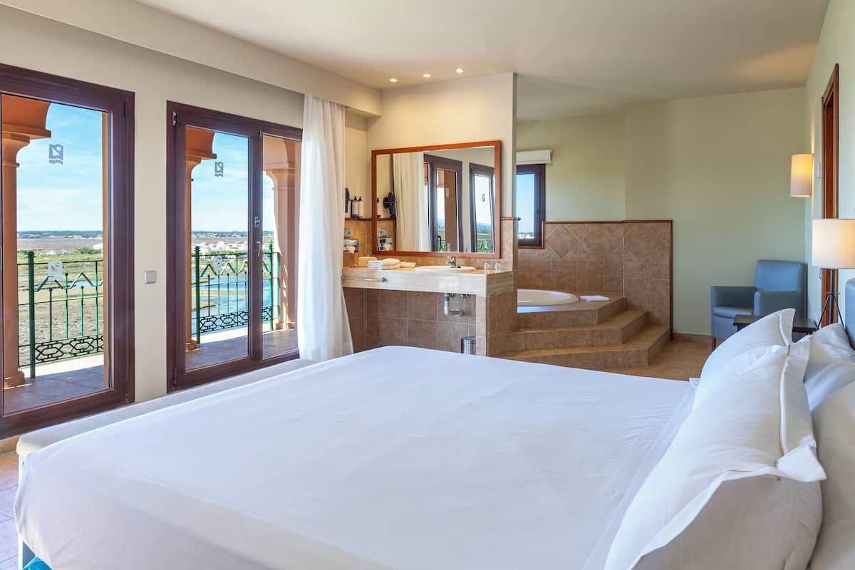 Une chambre d’hôtel avec un grand lit, une salle de bain avec un lavabo et un miroir, et un balcon avec vue.