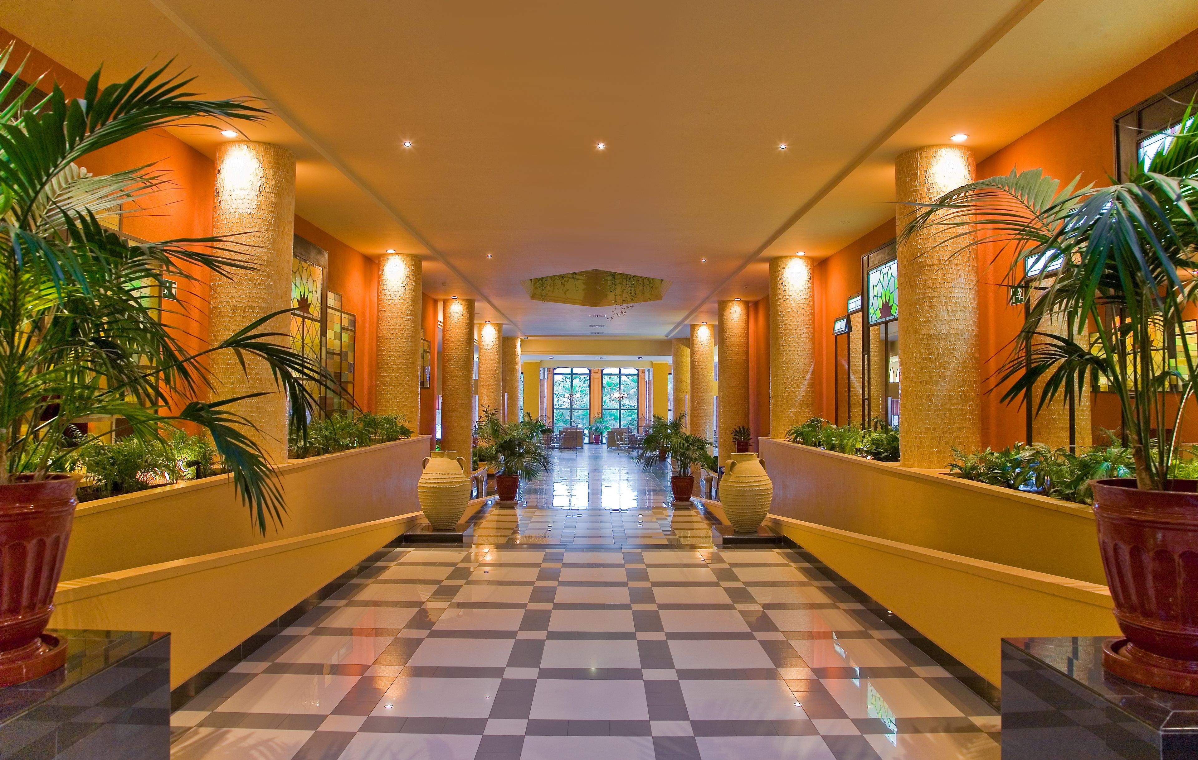 Ein Hotelkorridor mit Schachbrettfußboden, großen Kübelpflanzen und einer zentralen Leuchte.