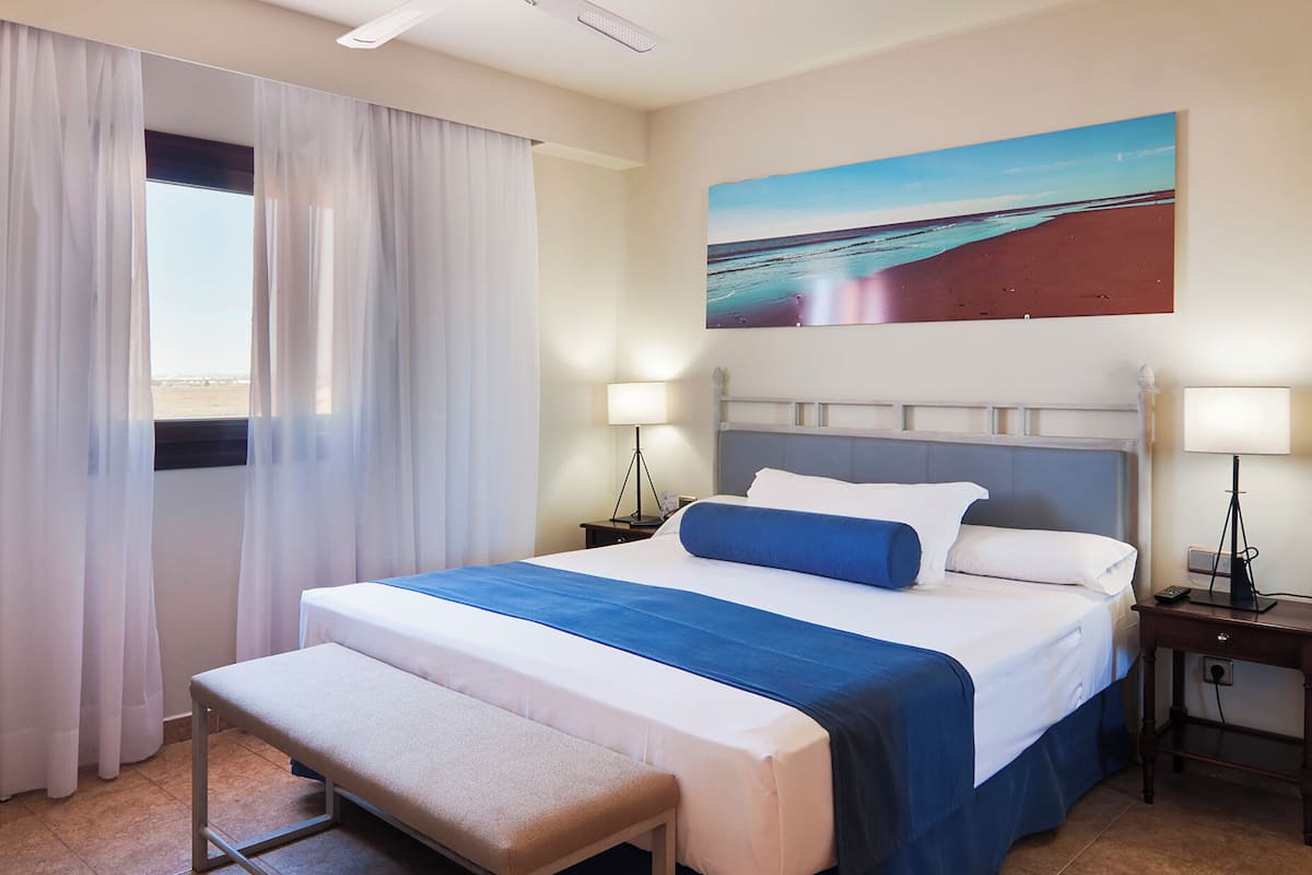 Une chambre d’hôtel avec un lit, une banquette, deux tables de chevet, un tableau et une fenêtre avec des rideaux.