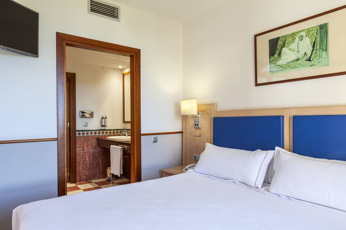 Une chambre d’hôtel avec un lit, une table de chevet, une lampe, un tableau et une salle de bain visible à travers une porte ouverte.