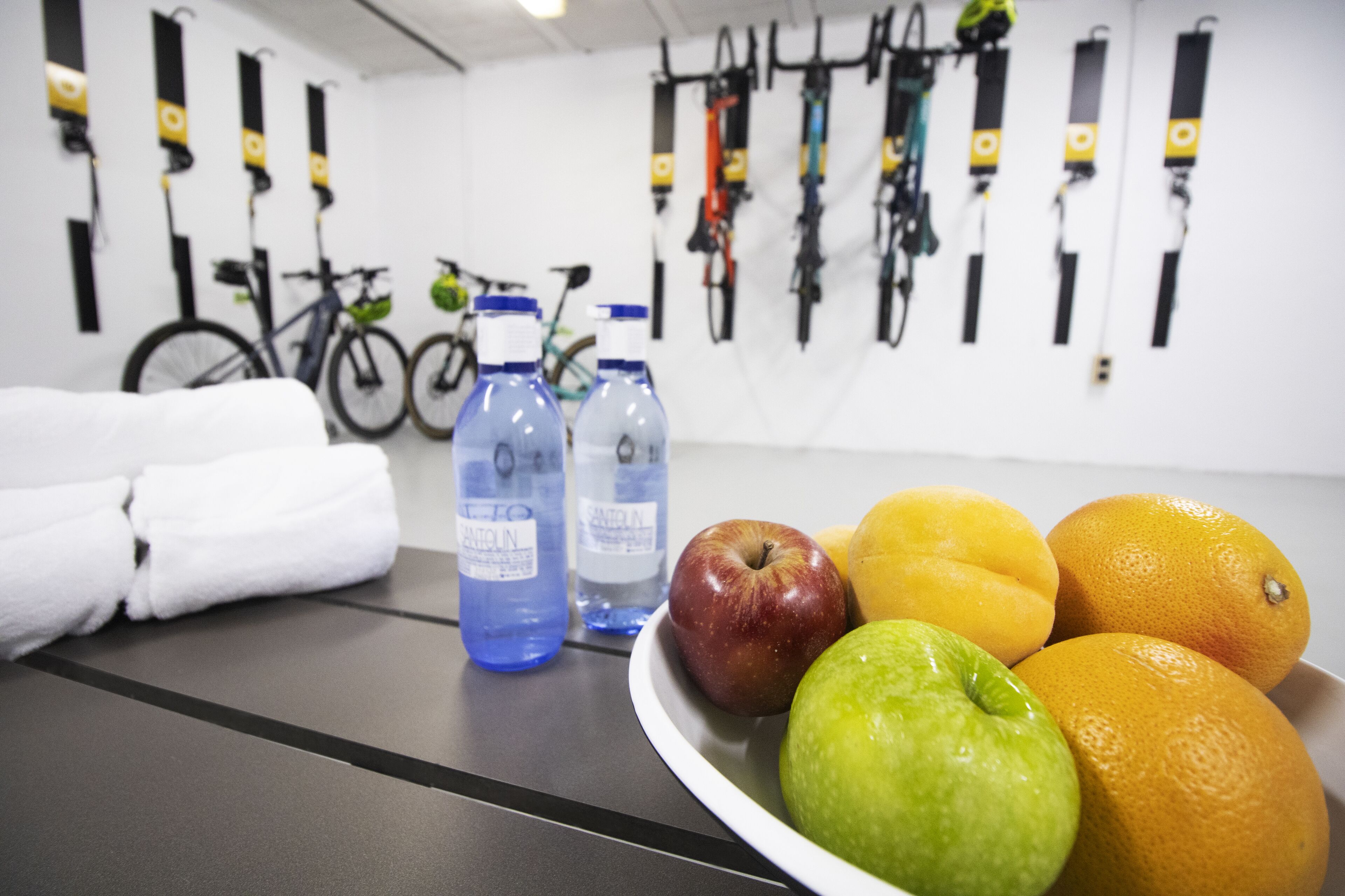 Une chambre avec un lit, une serviette, une bouteille d’eau, un panier de fruits et des vélos accrochés au mur.