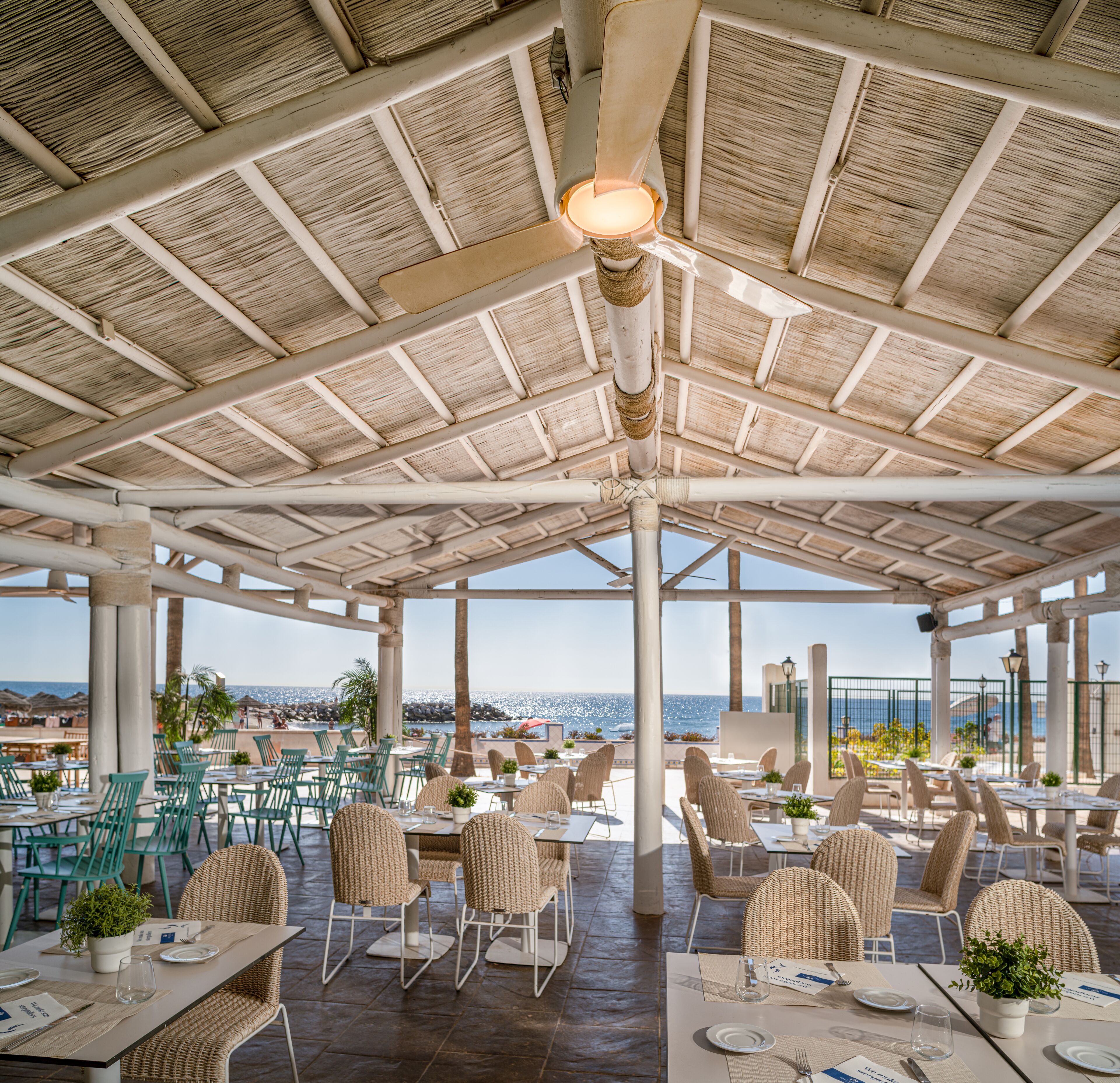 Un restaurante al aire libre con techo de paja, muebles de mimbre y vistas al mar.