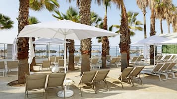 Un espace de détente extérieur, baigné de soleil, avec des rangées de chaises longues beiges et de parasols blancs, le tout entouré de grands palmiers.