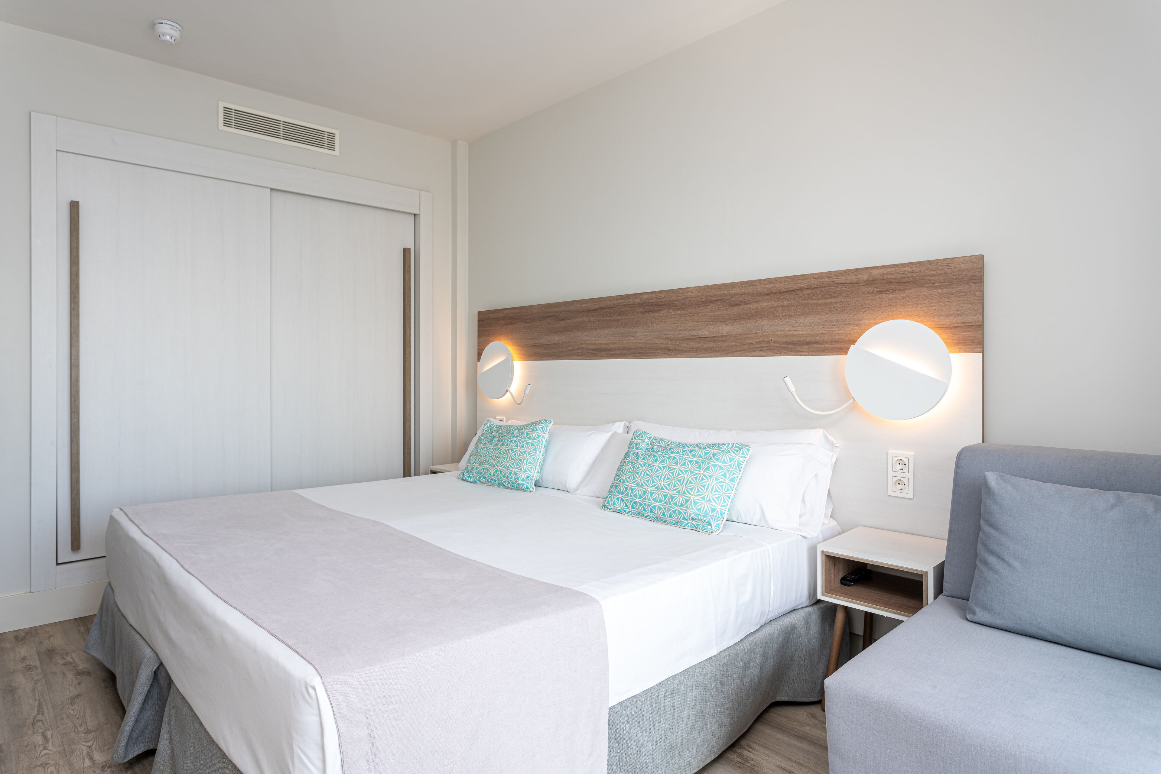 Een moderne hotelkamer met een groot bed, nachtkastjes, een grijze bank en een houten hoofdbord met ingebouwde verlichting.