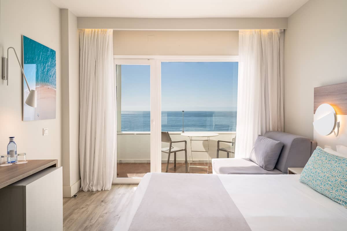 A hotel room with a balcony, a bed, a desk, and a view of the sea.