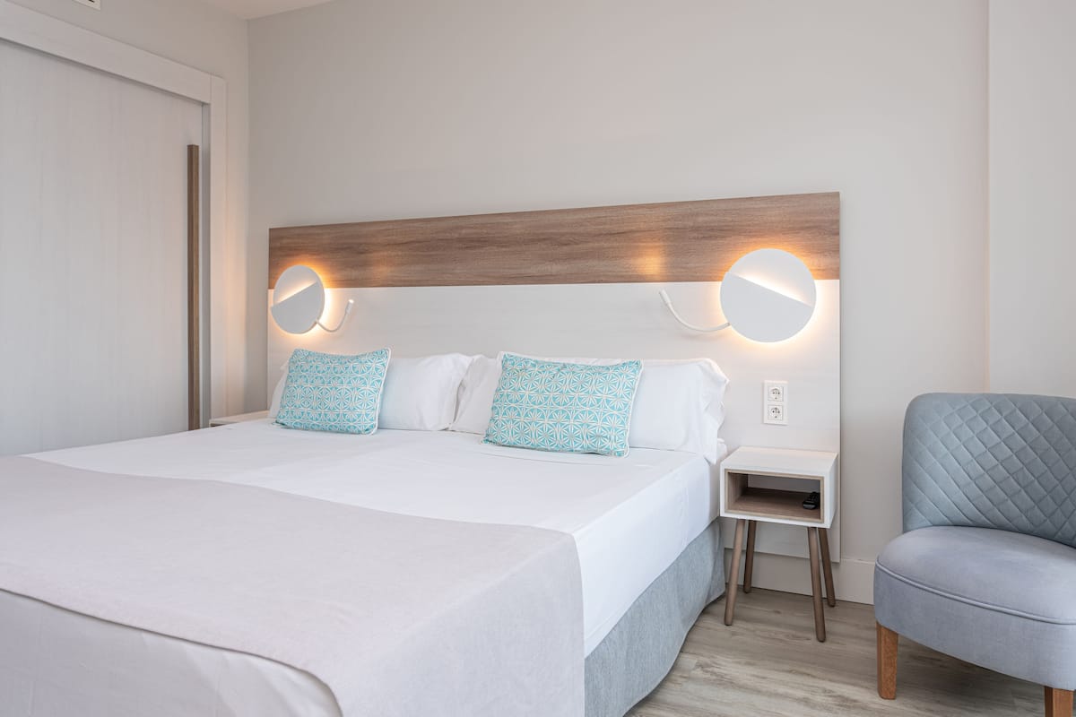 A neatly made bed with white bedding and patterned pillows, a bedside table with a lamp, and a blue upholstered chair.