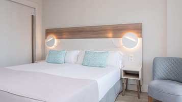 A neatly made bed with white bedding and patterned pillows, a bedside table with a lamp, and a blue upholstered chair.