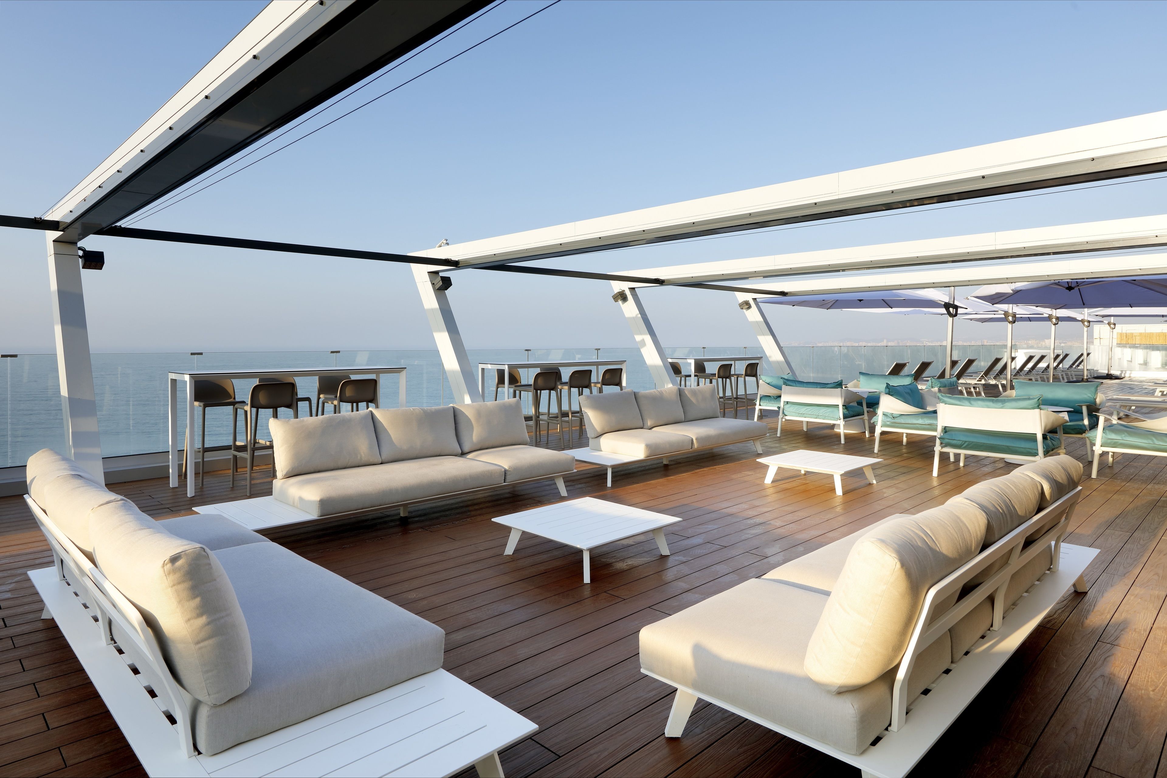 A sunlit outdoor deck with a wooden floor, white sofas, and a dining area with chairs and tables.