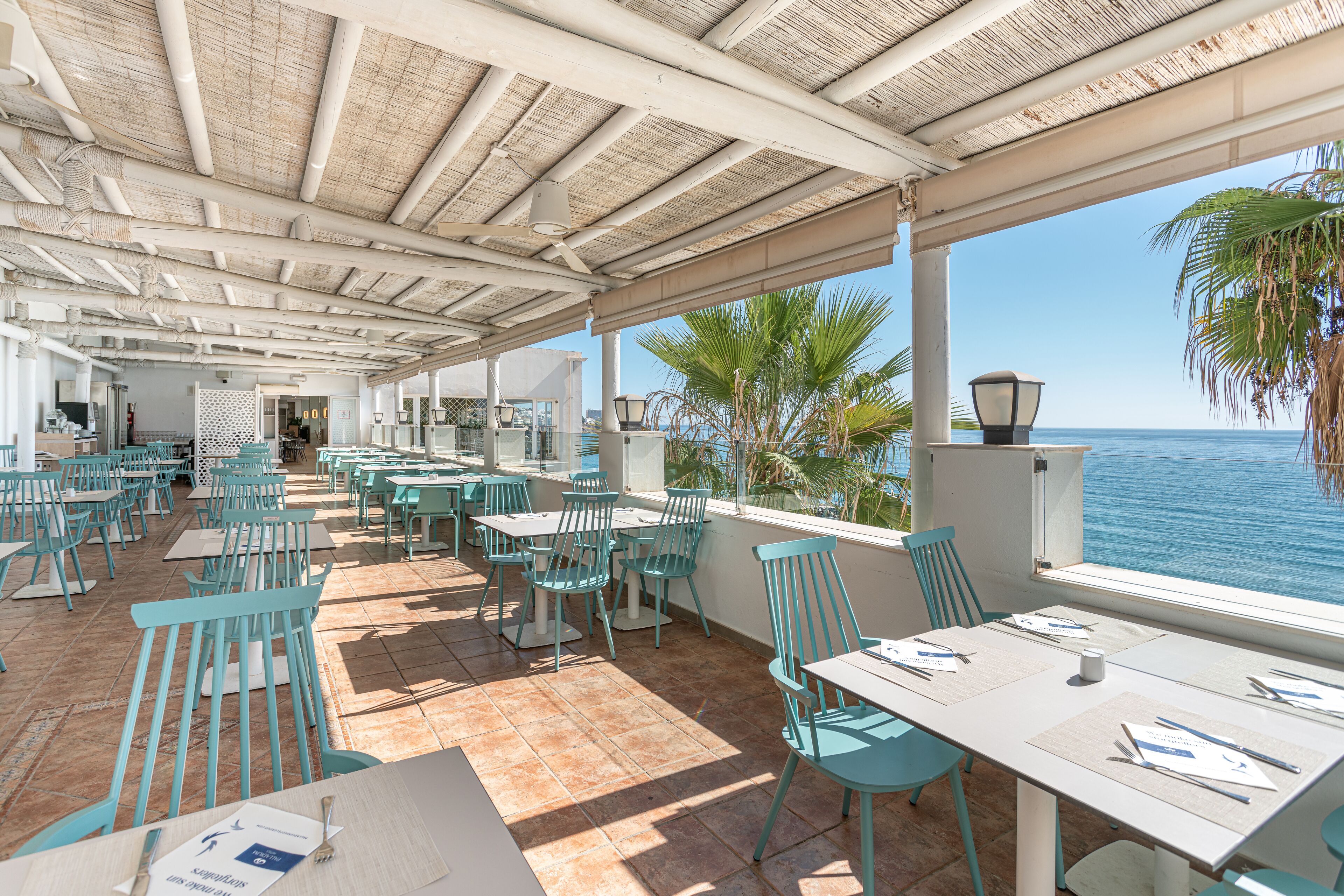 An outdoor dining area with tables and chairs, a view of the sea, and a covered roof structure.