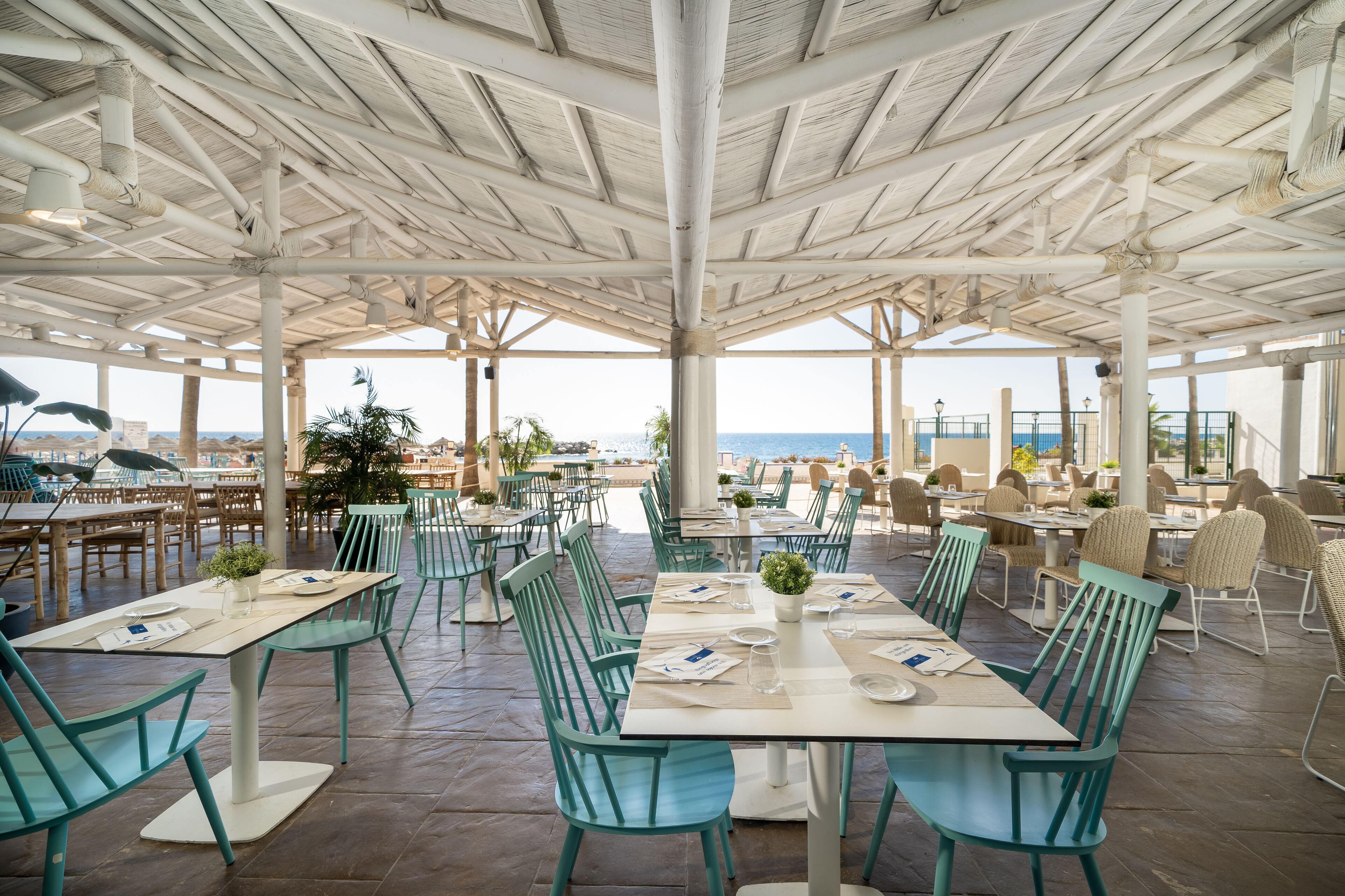 An outdoor dining area with tables and chairs, a view of the sea, and a covered structure with a white ceiling.