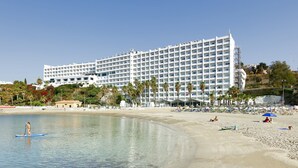 A large hotel with multiple floors and balconies, located near a beach with palm trees and beach umbrellas.