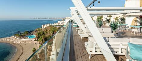 A balcony with a view of the beach and ocean, outdoor seating, and a pool area.
