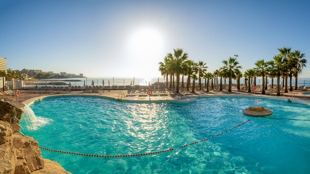 A swimming pool with clear blue water, surrounded by lounge chairs and palm trees, with a view of the ocean and buildings in the background.