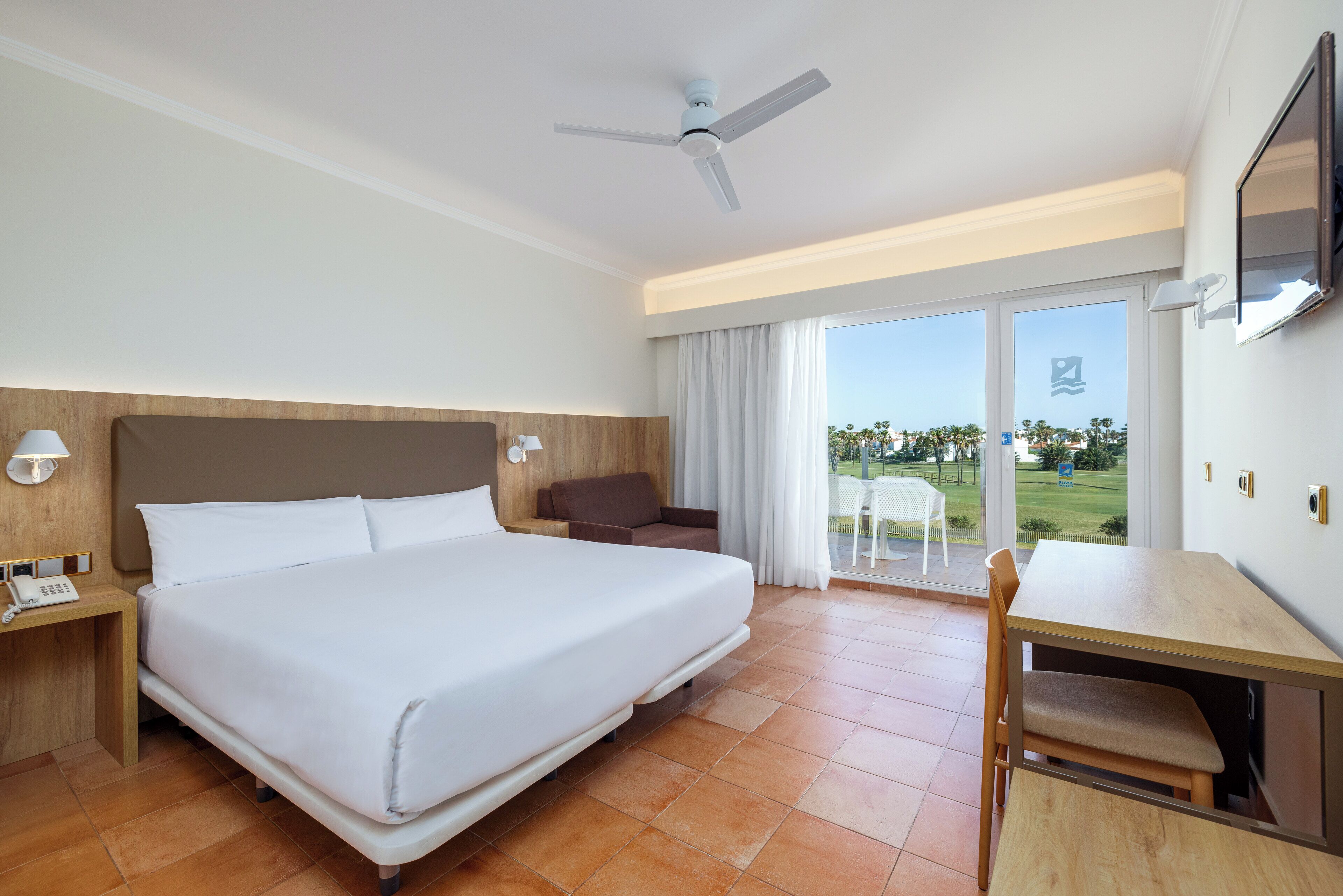 A hotel room with a large bed, a desk, a chair, a ceiling fan, and a view of the outdoors.
