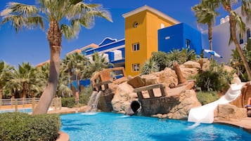 Una piscina con tobogán de agua, palmeras y un edificio con fachadas azules y amarillas.