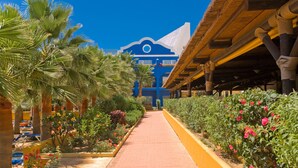 A pathway lined with palm trees and flowering plants leading to a building with a blue facade and a thatched roof structure. - Playaballena Hotel (Rota)