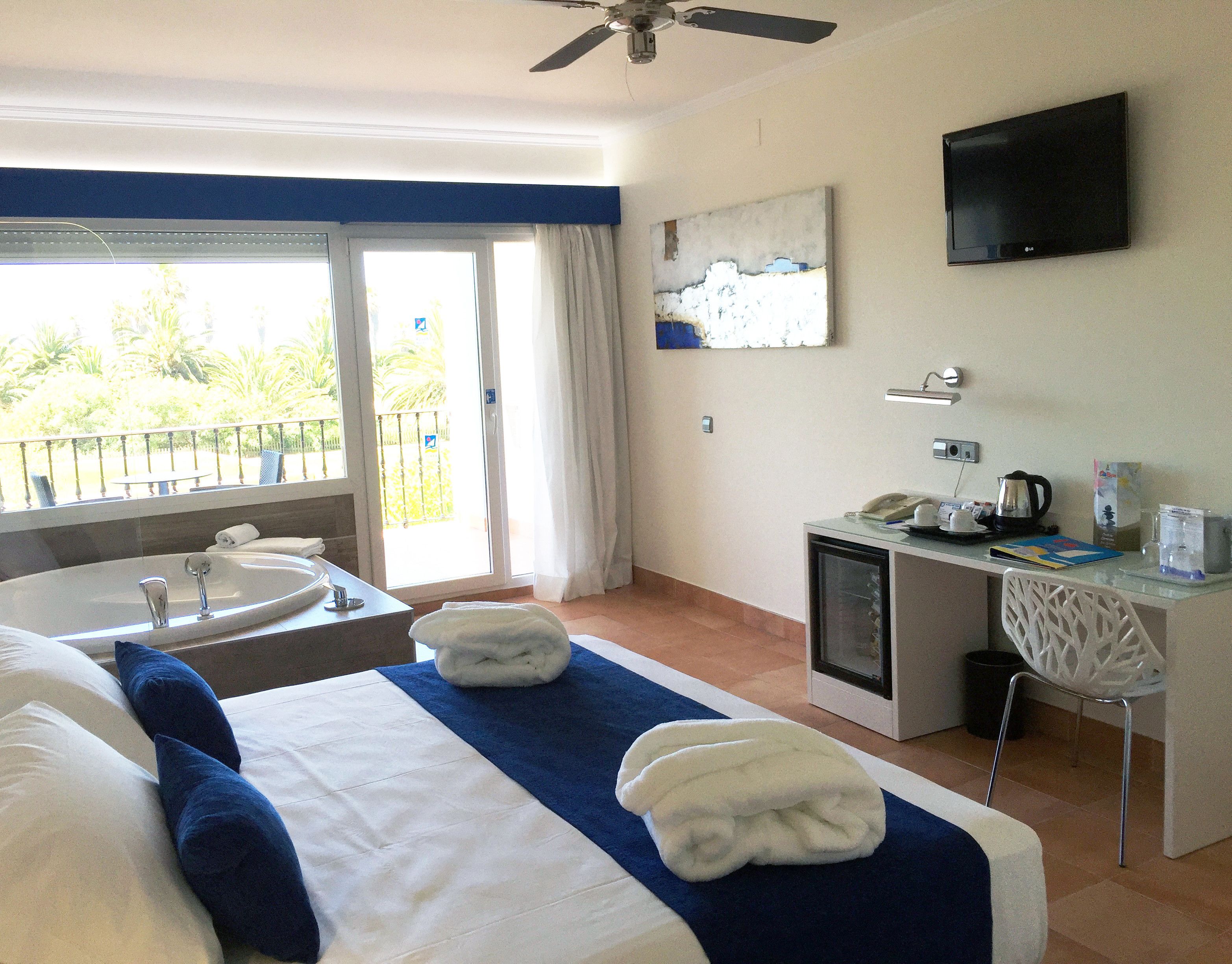 A hotel room with a bed, a desk, a chair, a TV, a window with a view of greenery, and a bathroom with a sink and mirror.