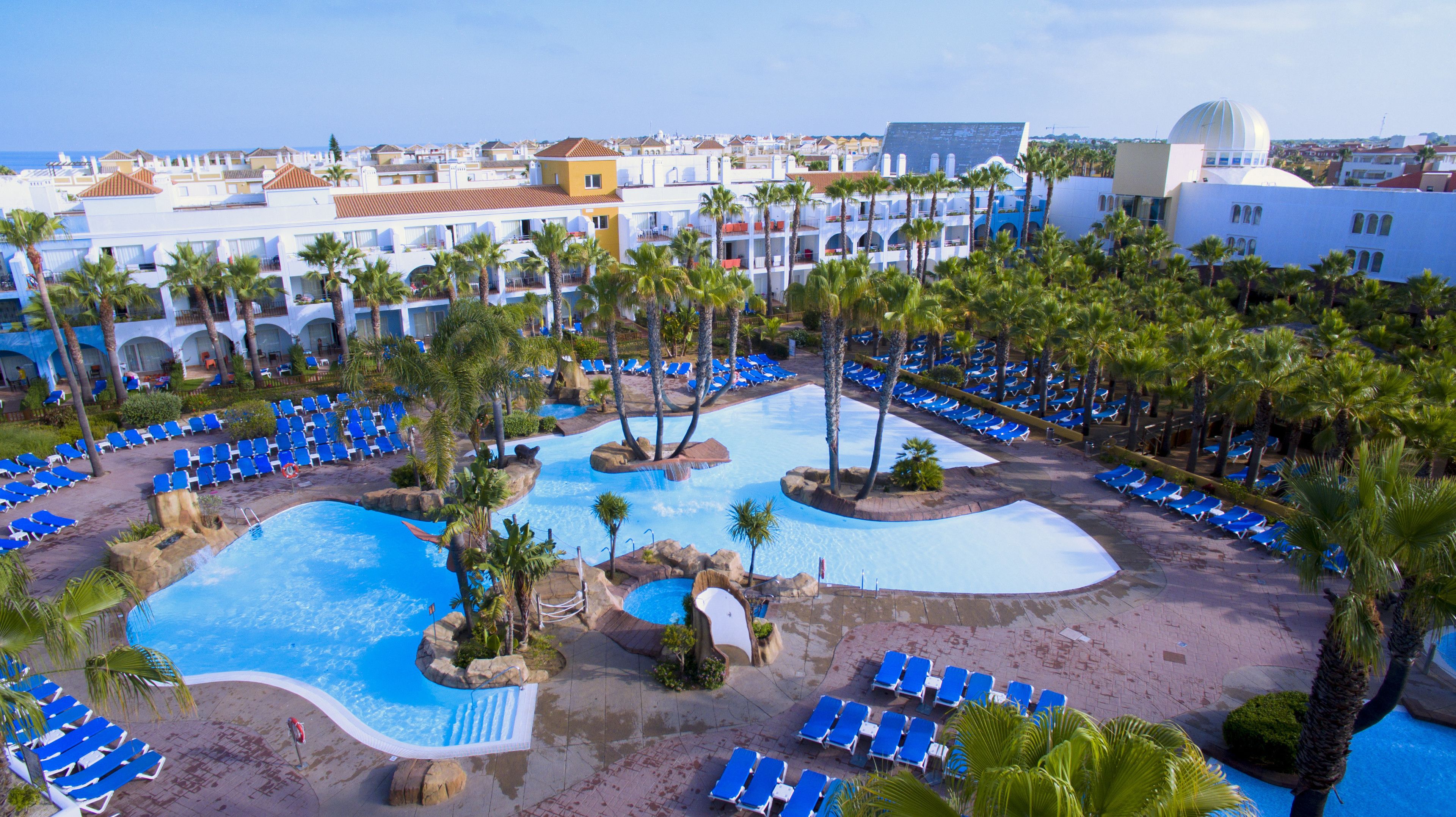 Un resort con varias piscinas, palmeras y sillones de descanso azules.