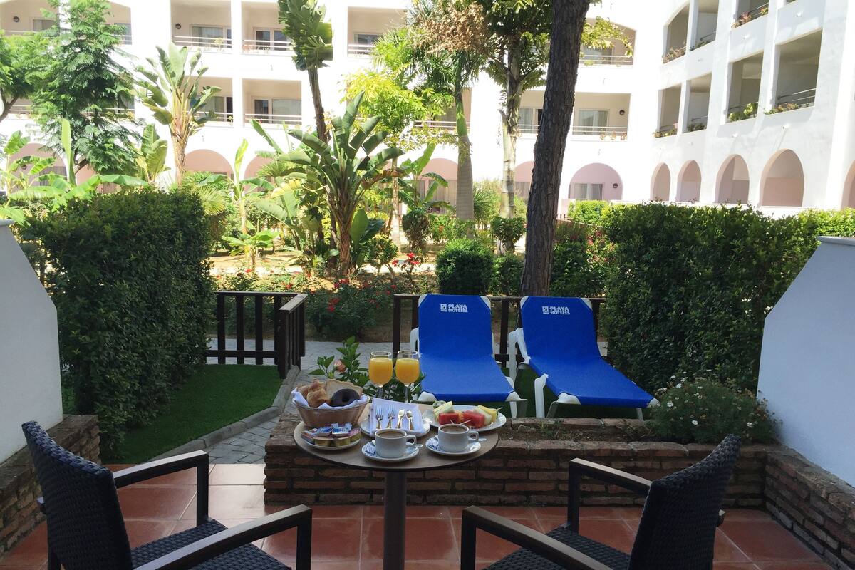 A patio with two blue lounge chairs, a table with breakfast items, and a view of a building with balconies.