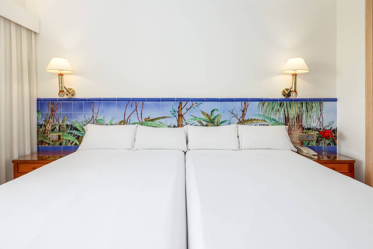A double bed with white linens, two bedside tables with lamps, and a mural of tropical plants on the headboard.