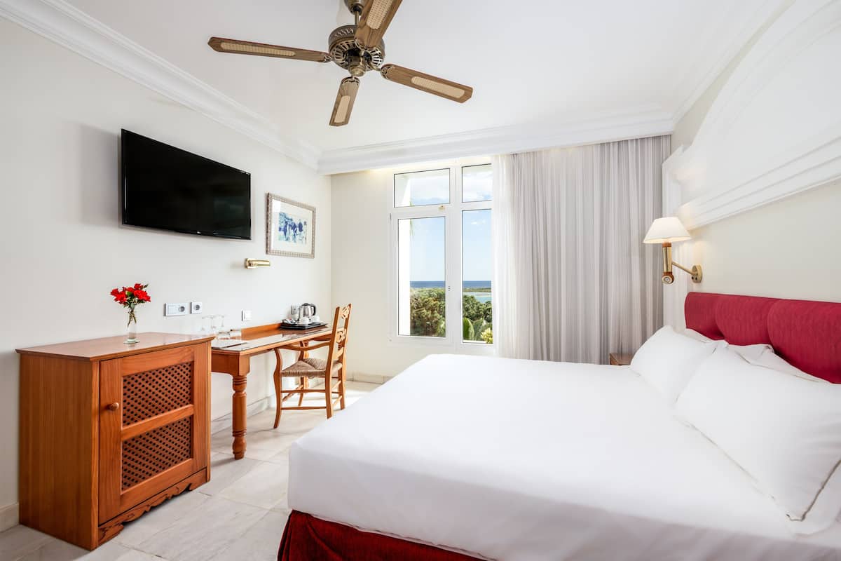 A hotel room with a bed, a desk, a television, a ceiling fan, and a window with curtains.