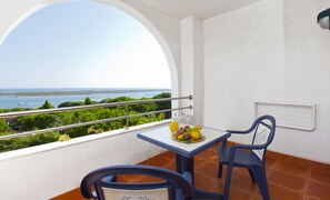 Un balcon avec une table dressée pour deux personnes, une chaise et une vue sur la mer et la verdure.