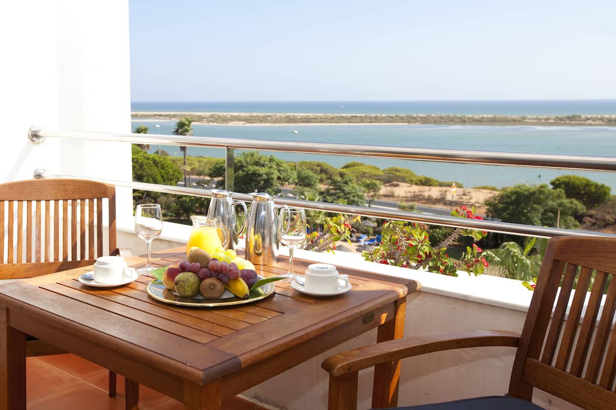 A wooden table with a fruit plate, two wine glasses, and a pitcher of orange juice on a balcony overlooking the sea.