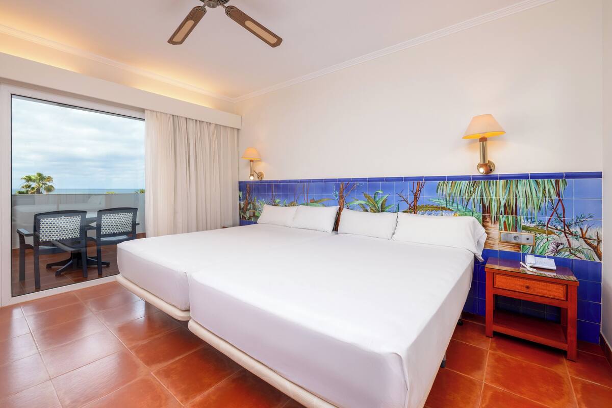 A hotel room with a large bed, two bedside lamps, a small table with a lamp, a chair, and a view of the ocean through a sliding door.