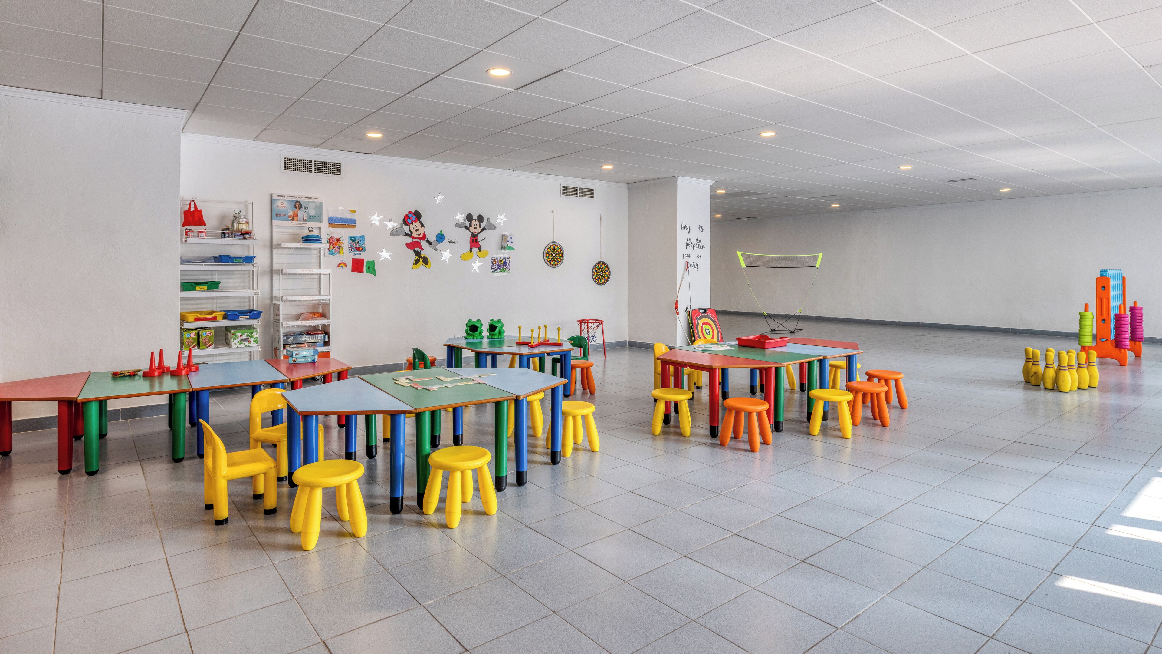 A brightly lit classroom with colorful tables and chairs, educational posters, and a variety of learning materials.