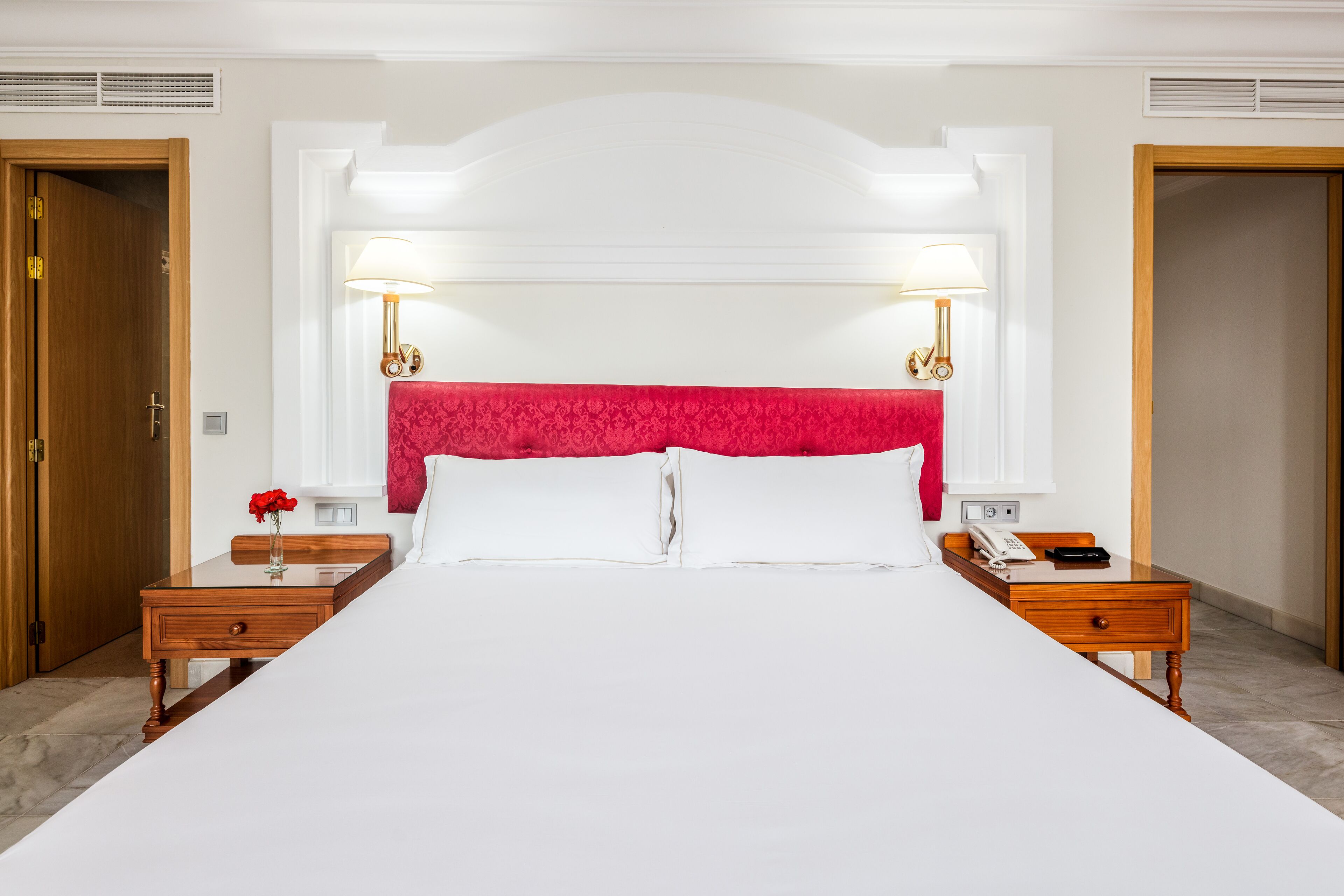 A hotel room with a large bed, two bedside tables, a red headboard, and wall-mounted lamps.