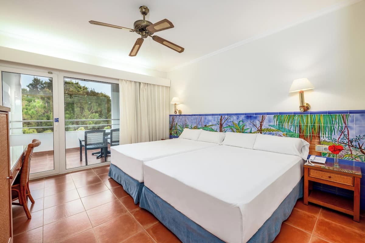 A hotel room with a large bed, a ceiling fan, a balcony with a chair, and a decorative wall mural.