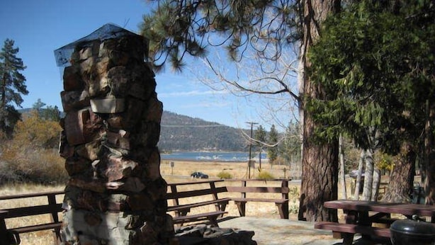 A stone structure with a metal roof, surrounded by picnic tables and a barbecue grill, overlooking a beach and hills. - Timberline Lodge (Big Bear Lake)
