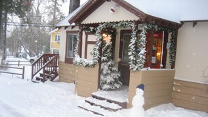 A snow-covered house with a wooden staircase leading to the front door, a small porch with a Christmas wreath, and a snowman figure in front. - Timberline Lodge (Big Bear Lake)