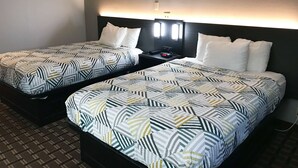 A hotel room with two beds, each with a patterned bedspread, a bedside table with a clock, and a wall-mounted light fixture. - Studio 6 Suites Tupelo, MS - North (Tupelo)