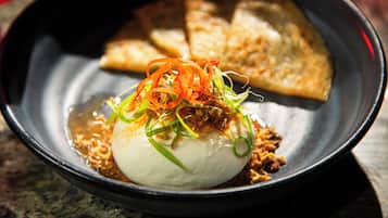 A bowl of food with a white spherical item topped with vegetables and sauce, accompanied by two pieces of bread.