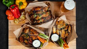 Una mesa de madera con tres cestas de papel que contienen alitas de pollo, apio, zanahorias y dos salsas para mojar.