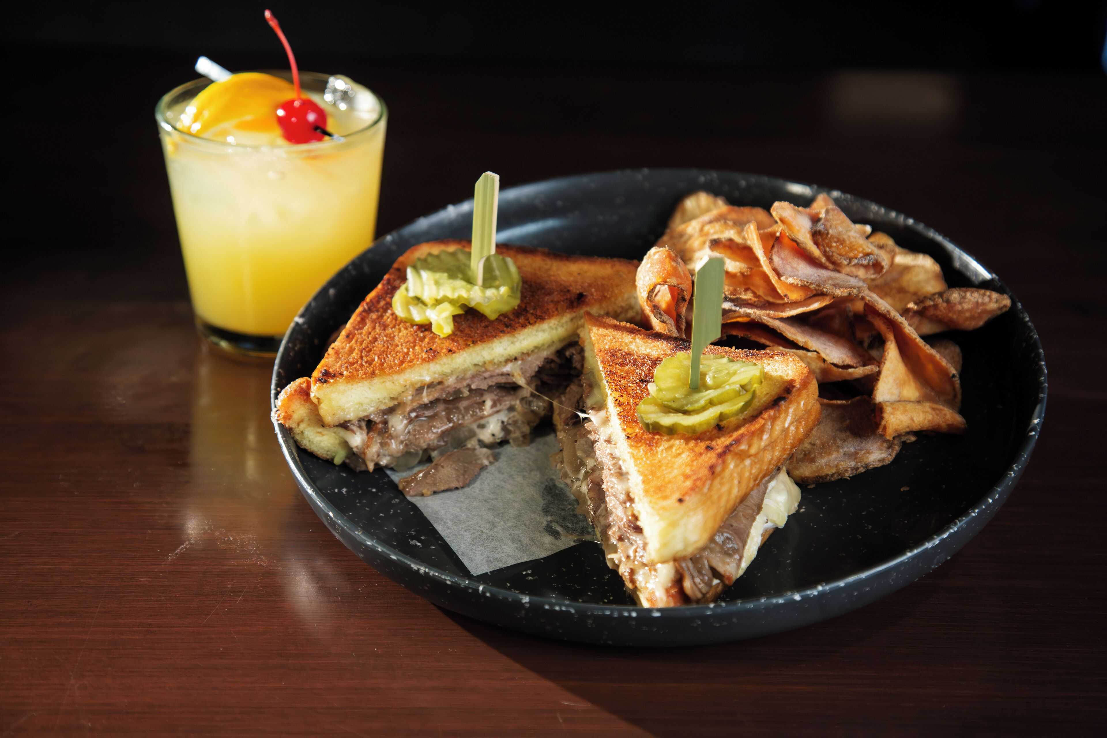 A plate with a grilled sandwich and fried food, and a glass with a yellow drink and a cherry.