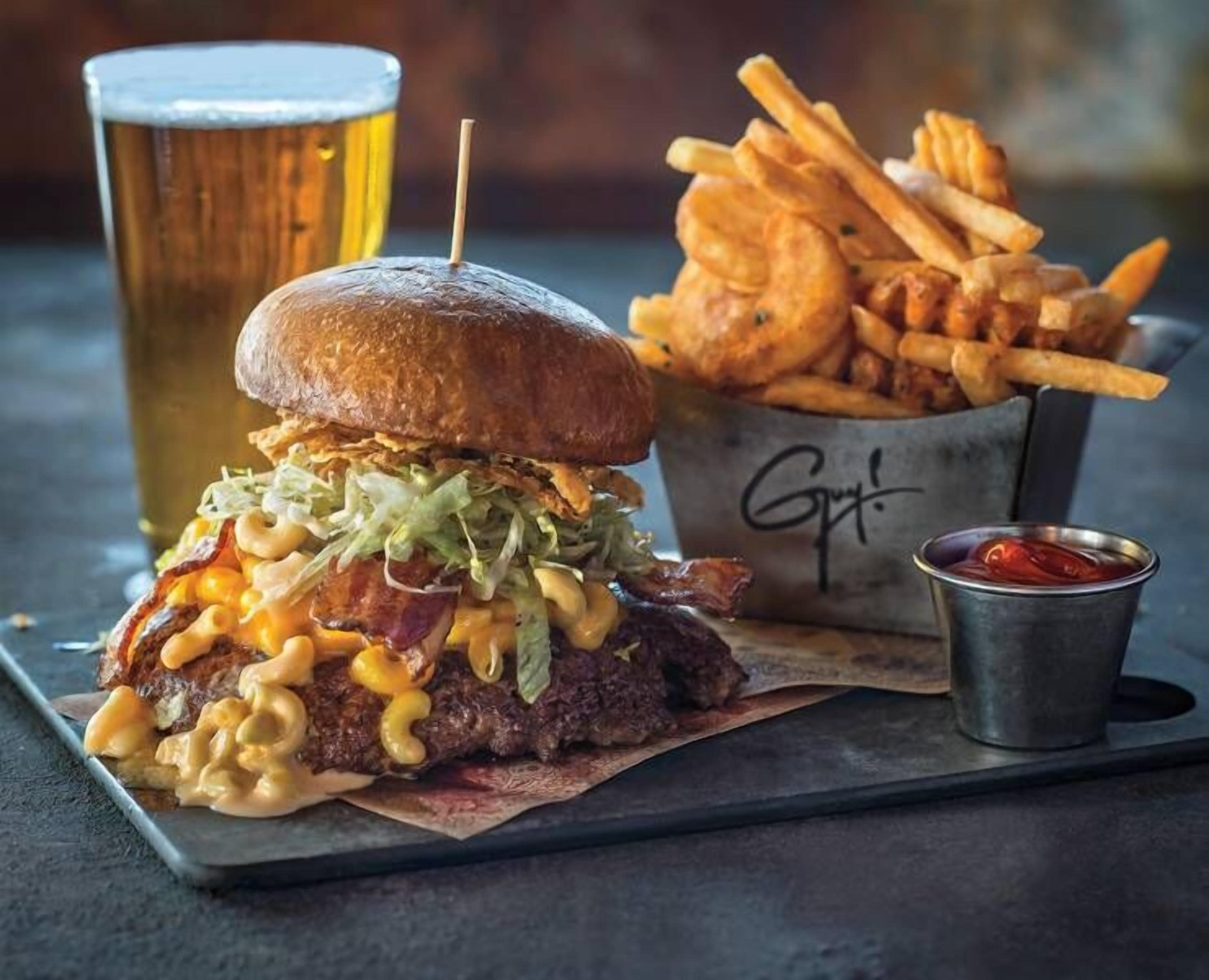 A burger with mac and cheese, bacon, and lettuce on a bun, served with fries and a cup of beer.