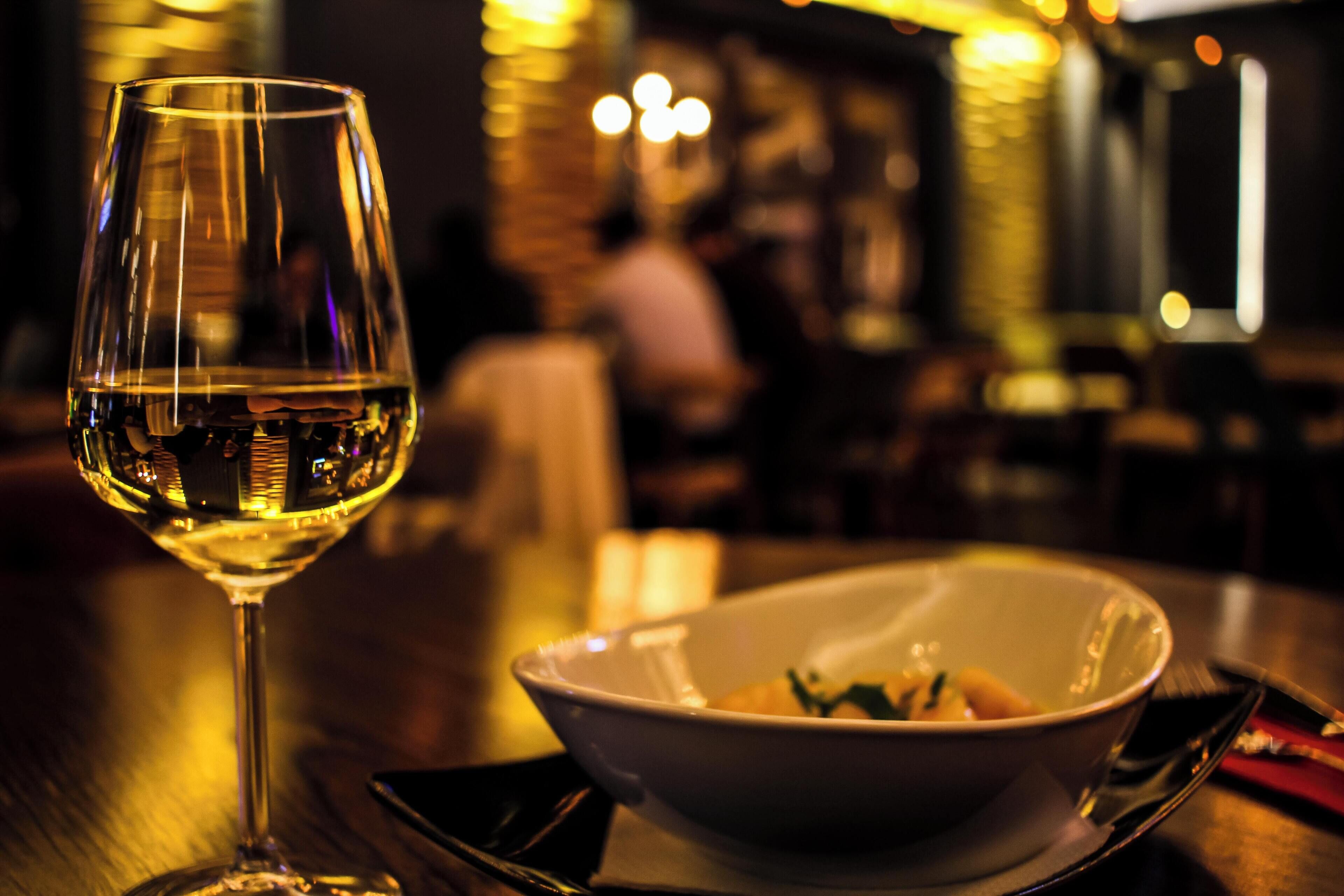 Una copa de vino blanco sobre una mesa con un bol de comida y cubiertos.