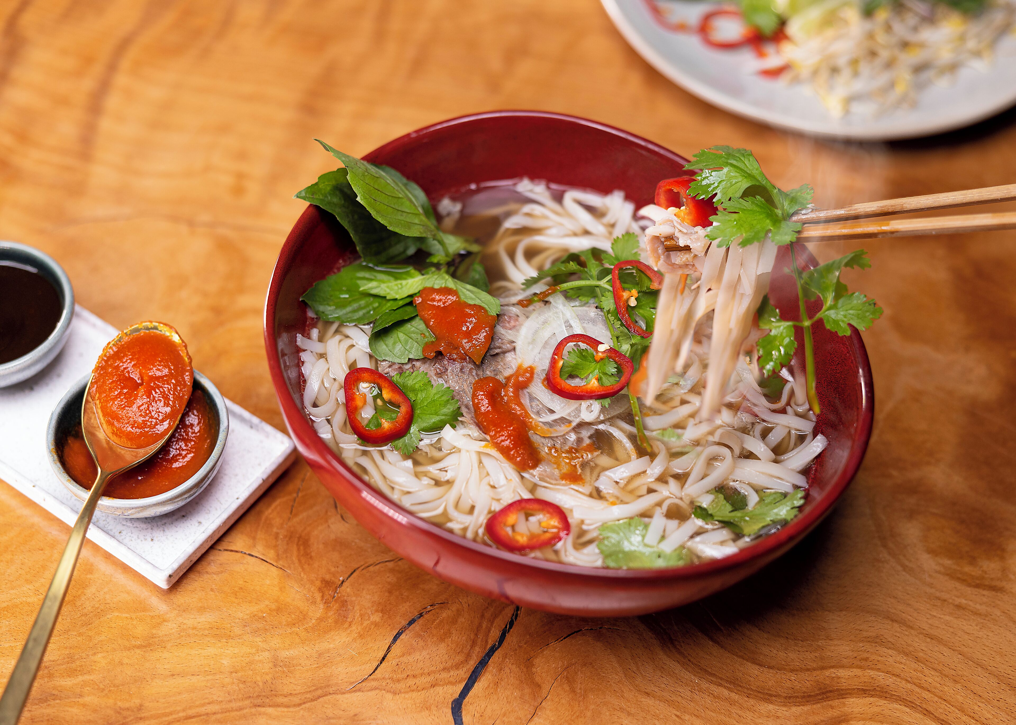 A bowl of noodle soup with various toppings, a spoon with sauce, and a pair of chopsticks.