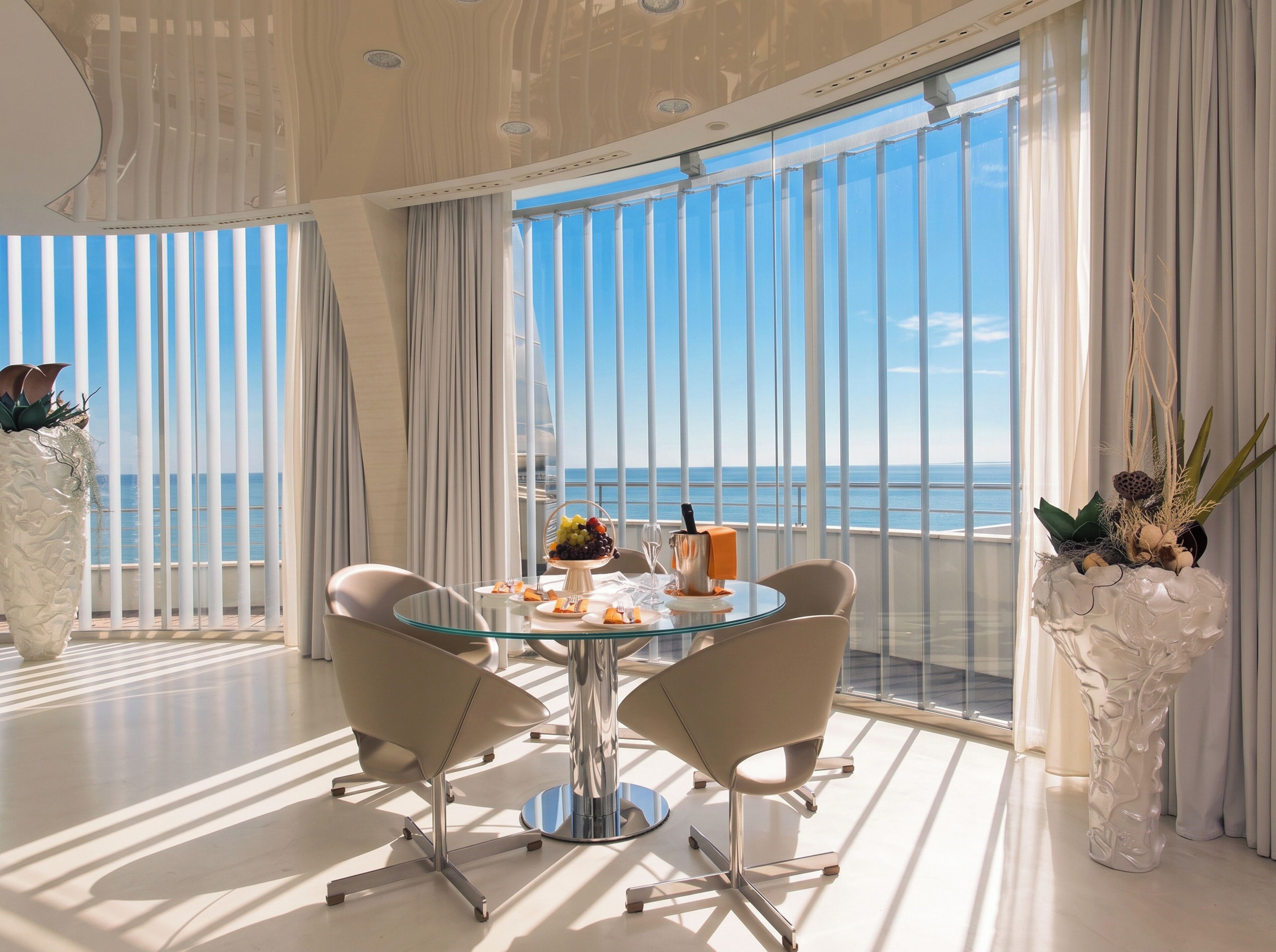 A modern dining area with a glass table, two chairs, and a view of the sea through large windows.