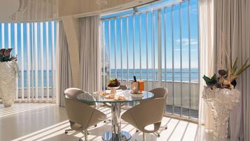 A modern dining area with a glass table, two chairs, and a view of the sea through large windows.
