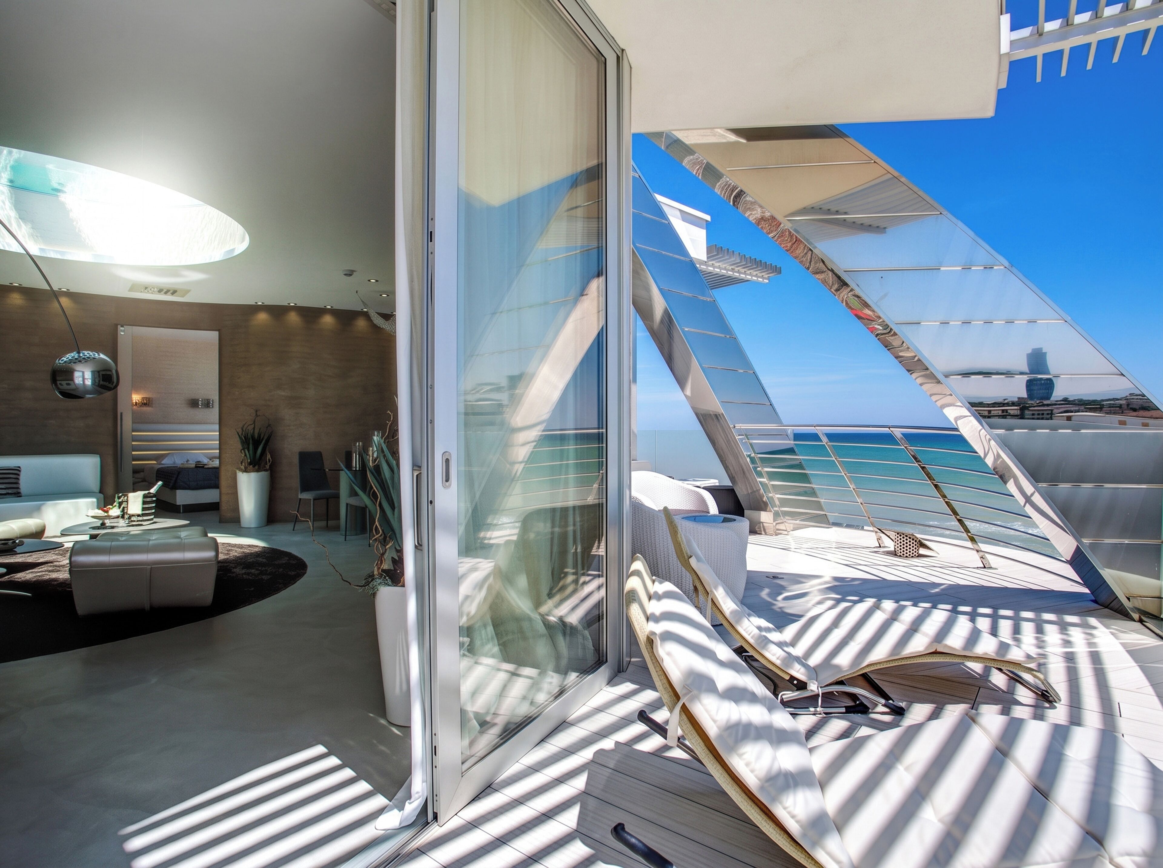 A modern interior with a glass door leading to a balcony with sun loungers, a sofa, and a dining area.