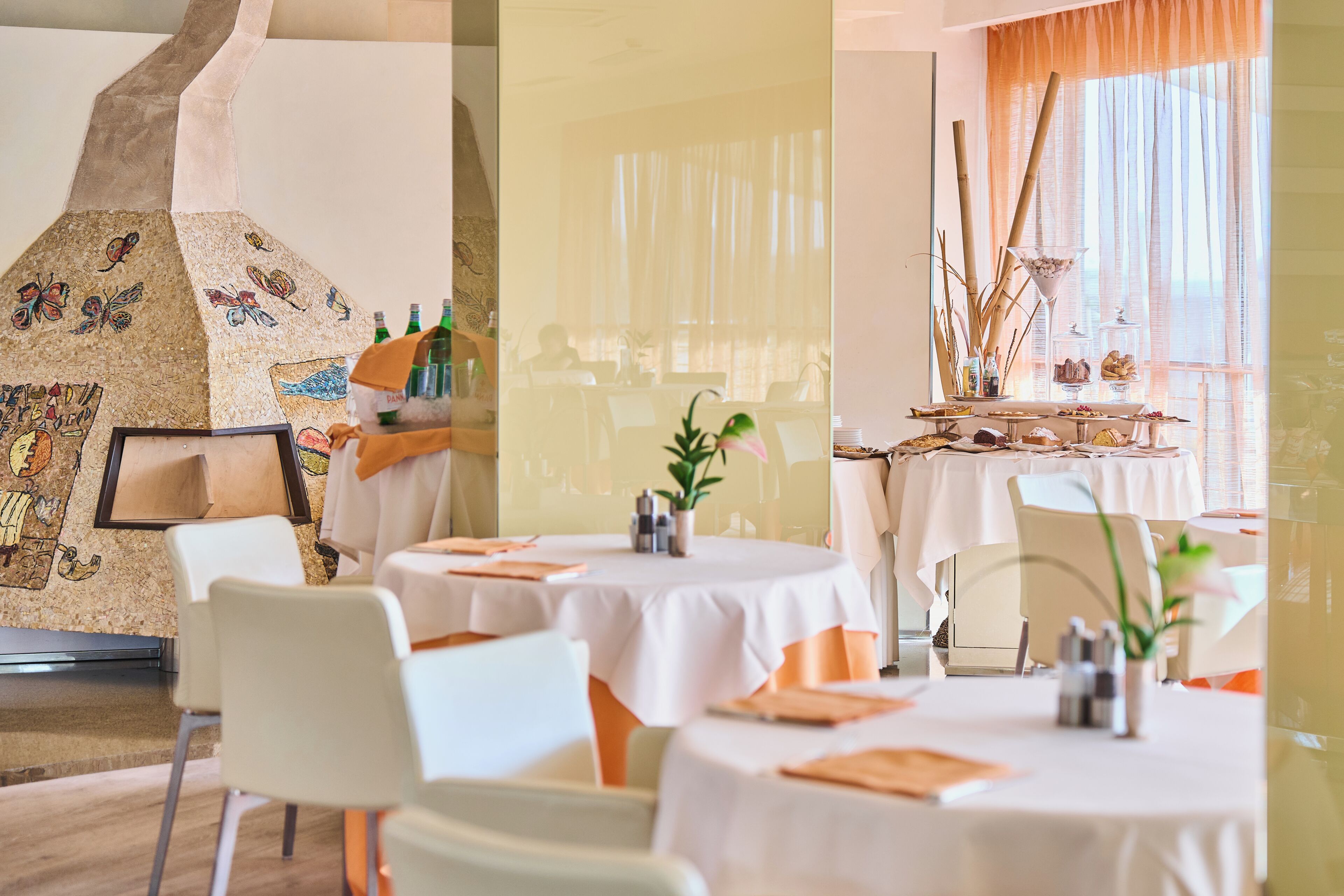Un restaurante con mesas redondas, sillas blancas y una pared decorativa con mosaicos.