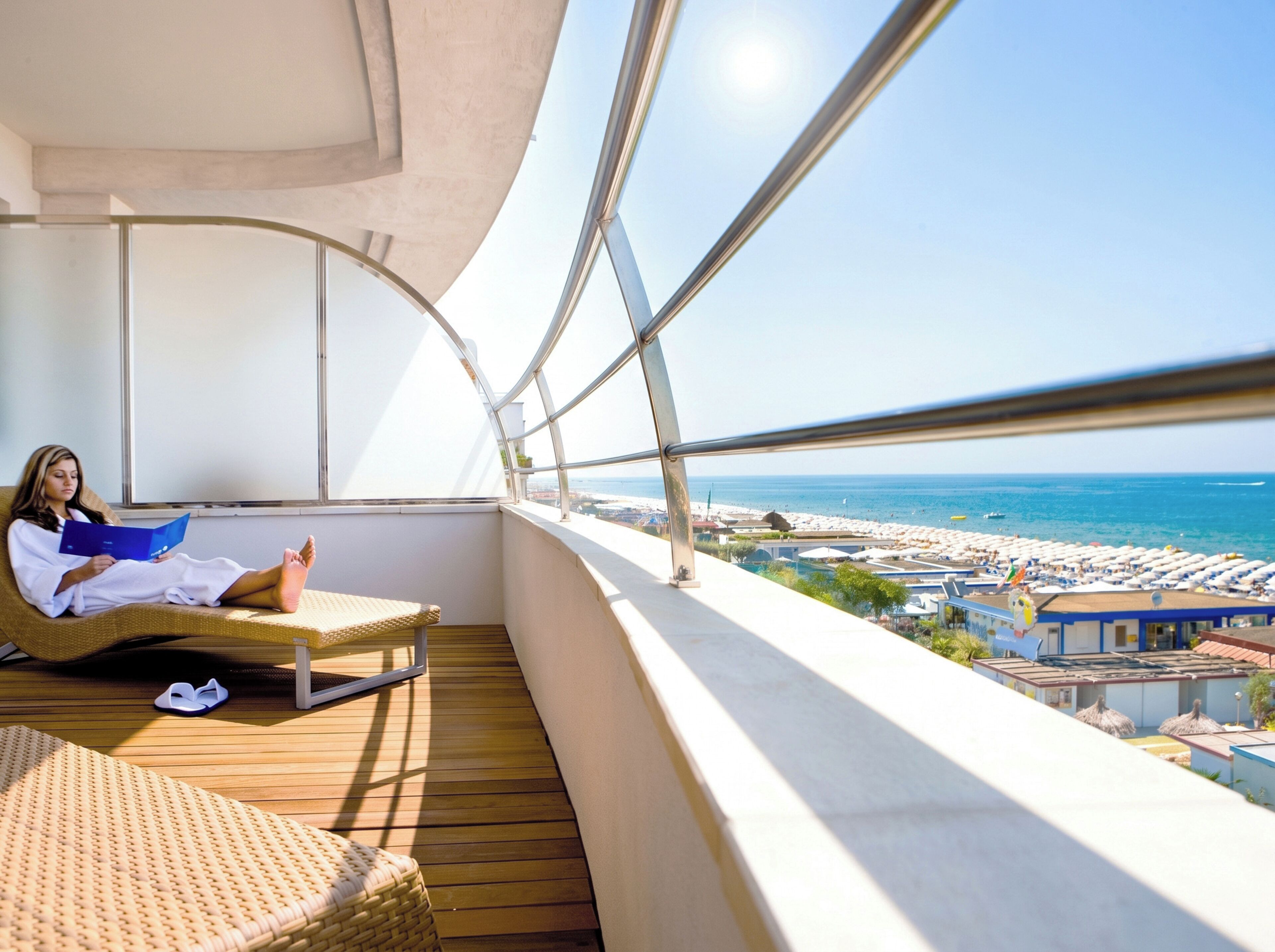 Un balcone con vista sulla spiaggia e sul mare, una persona che legge e una sedia in vimini.
