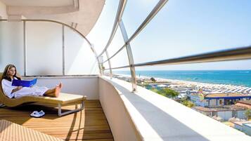 Un balcone con vista sulla spiaggia e sul mare, una persona che legge e una sedia in vimini.