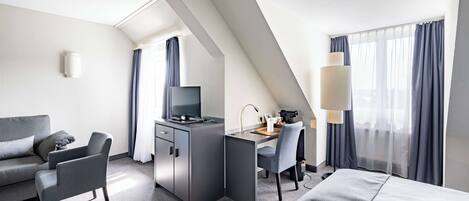 Une chambre d’hôtel moderne équipée d’un canapé, d’un bureau avec un ordinateur, d’un lit et d’une grande fenêtre avec des rideaux.