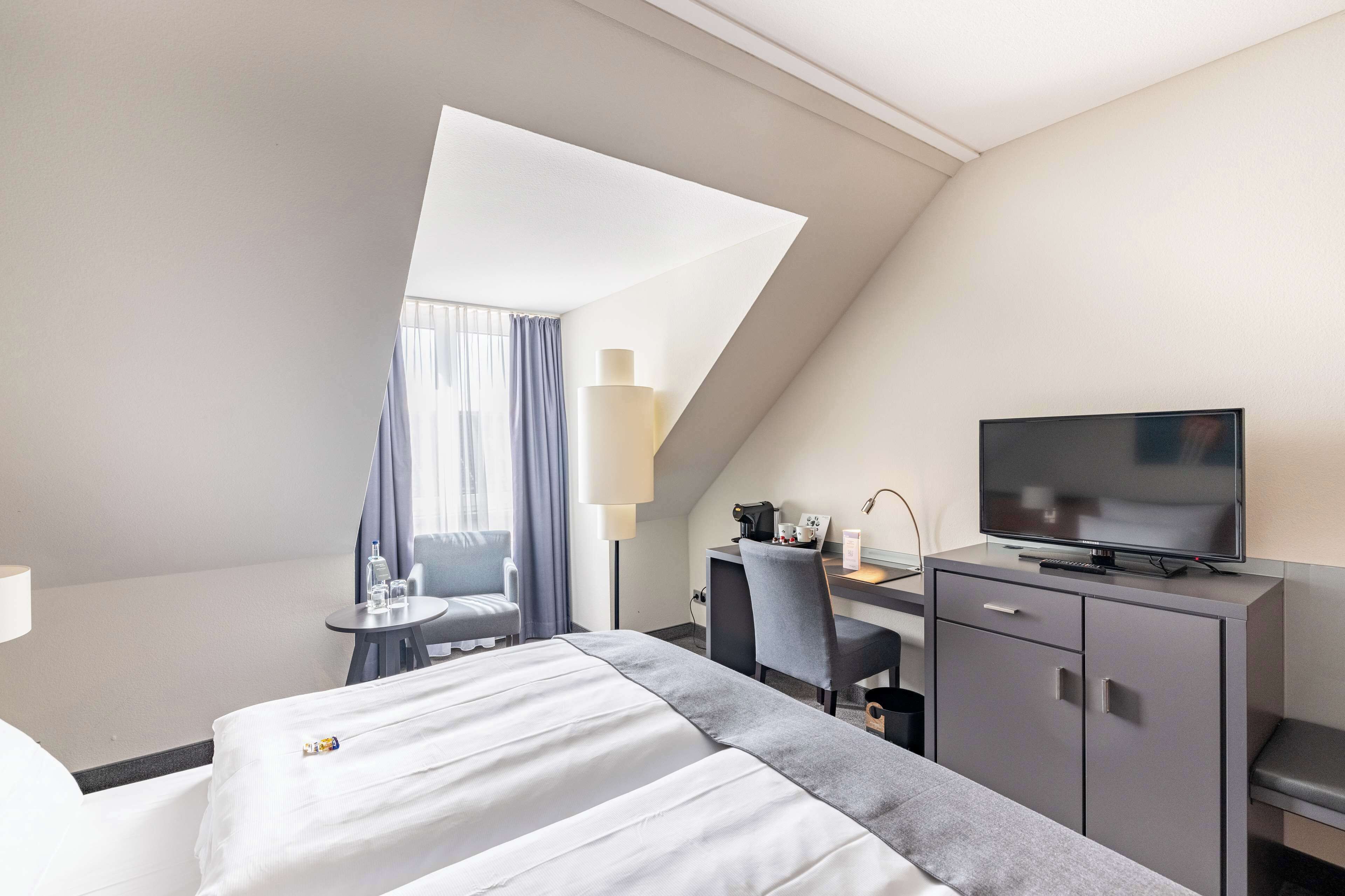 Ein Hotelzimmer mit einem Bett, einem Schreibtisch mit Computer, einem Fernseher und einem kleinen Tisch mit Lampe.