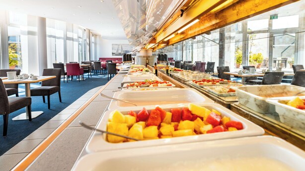 A buffet area with various food options, tables, and chairs arranged in a spacious room with large windows. - Atlantic Hotel Vegesack (Bremen)