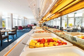 A buffet area with various food options, tables, and chairs arranged in a spacious room with large windows. - Atlantic Hotel Vegesack (Bremen)